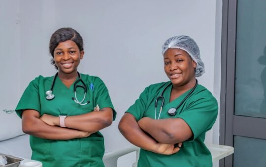 Two female nurses in green scrubs smiling confidently in a hospital room. Professional healthcare workers.