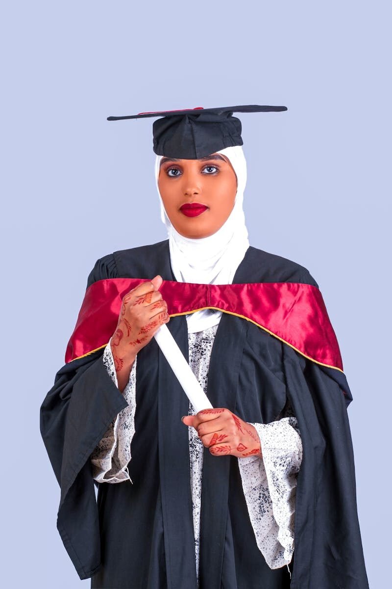 Confident graduate woman in cap and gown holding diploma against a neutral background.